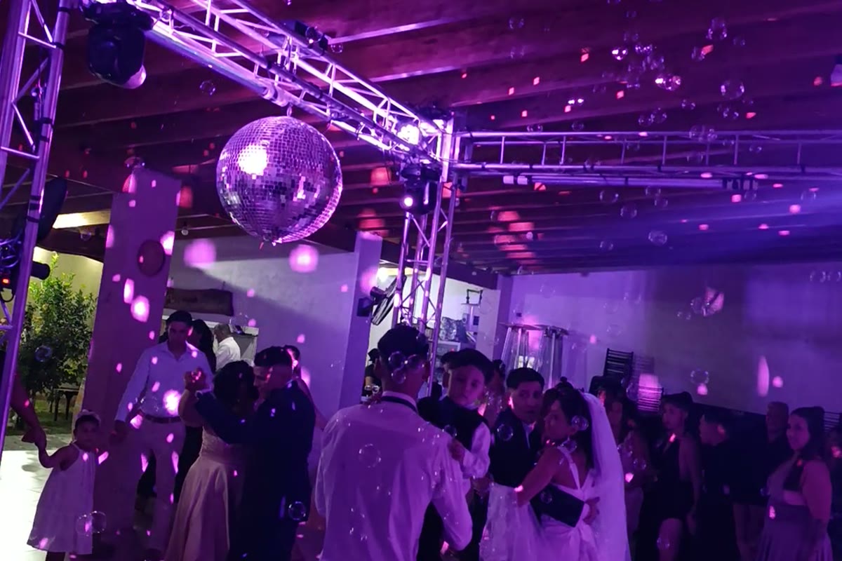 El mismo salón a la medianoche: luz violeta, bola de espejos encendida, burbujas y la pista llena de invitados bailando